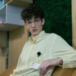 Young man in a yellow shirt with ID badge sitting in a modern office setting.